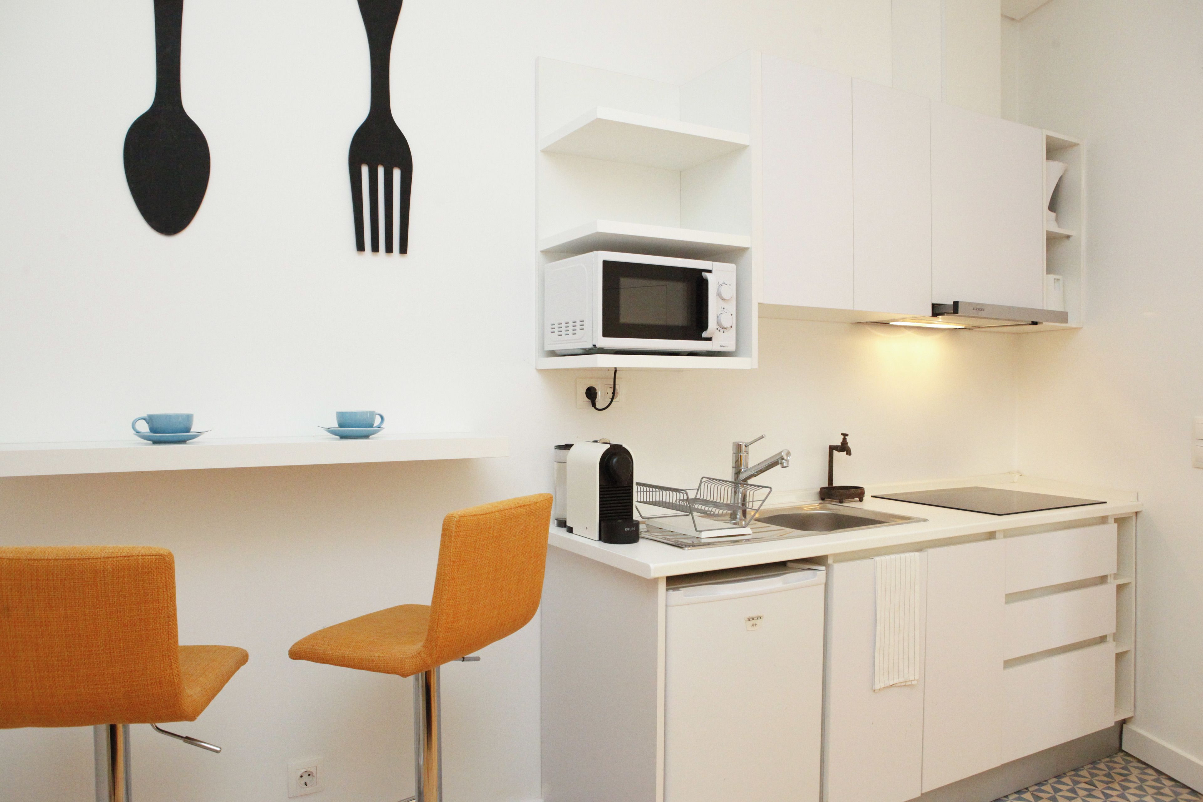 A compact kitchen with white cabinets, a microwave, a sink, and two orange chairs.