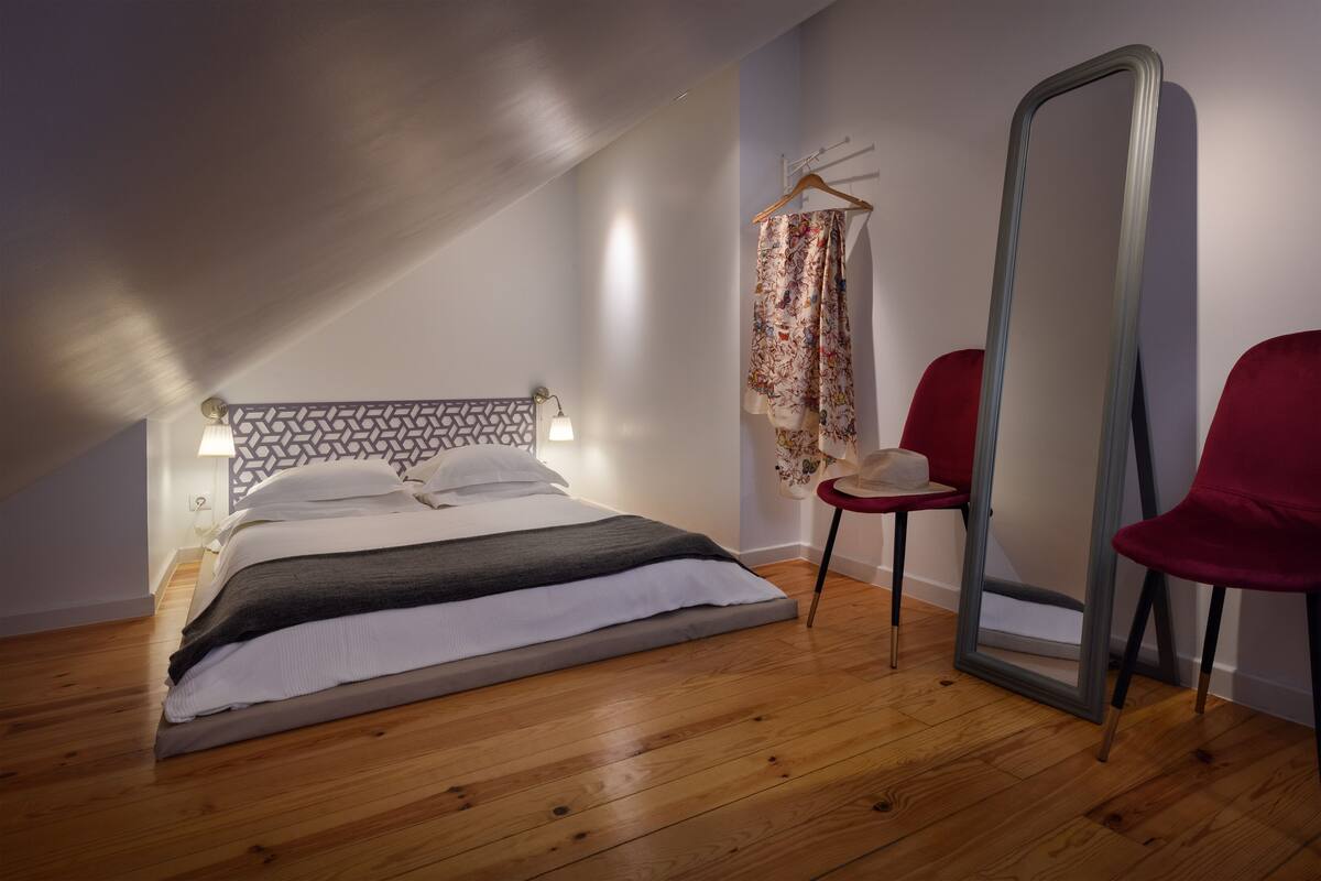 A bedroom with a wooden floor, a bed with white and gray bedding, a hanging garment, a mirror, and a red chair.