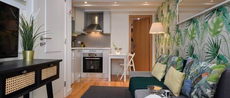A compact living space with a kitchenette, a sofa with cushions, a small table, and a potted plant.