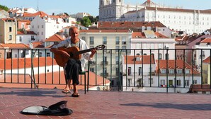 Um homem toca violão em um terraço, com uma paisagem urbana e uma catedral imponente ao fundo.
