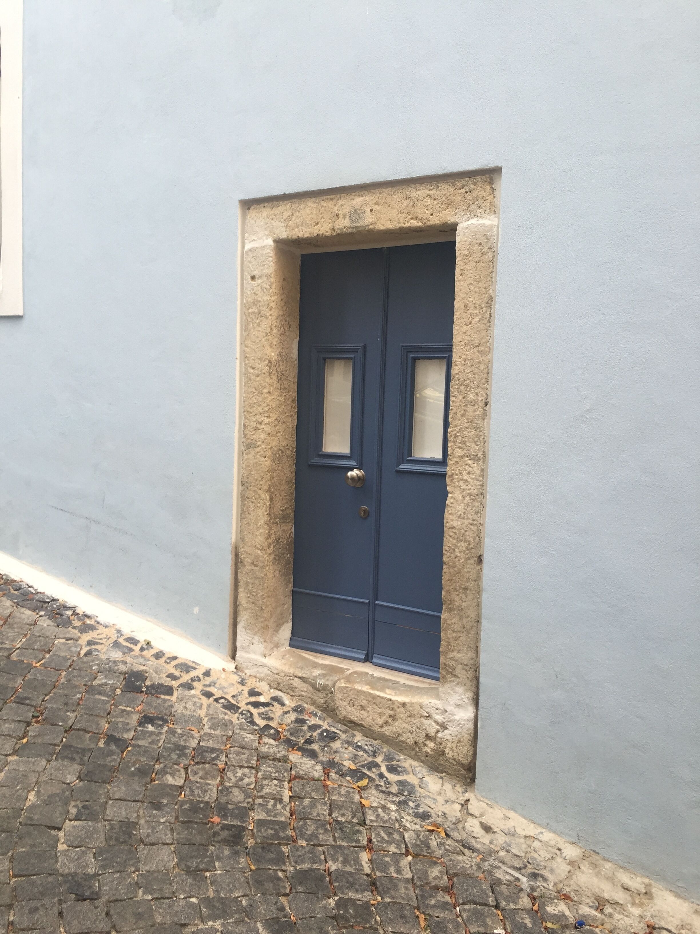 Eine blaue Tür mit kleinem Fenster, in ein Steingemäuer eingesetzt, vor einer hellblauen Wand.