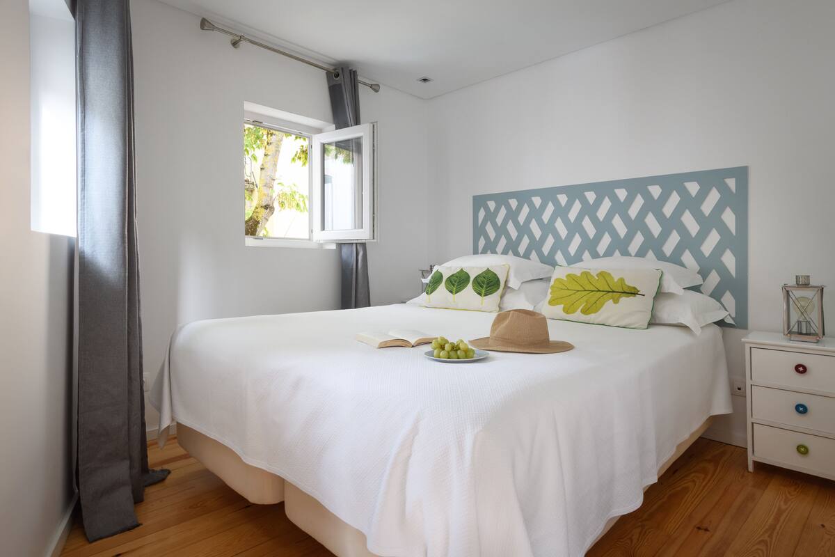A neatly made bed with a white bedspread, a window with a view of greenery, and a bedside table with a lamp and a decorative item.