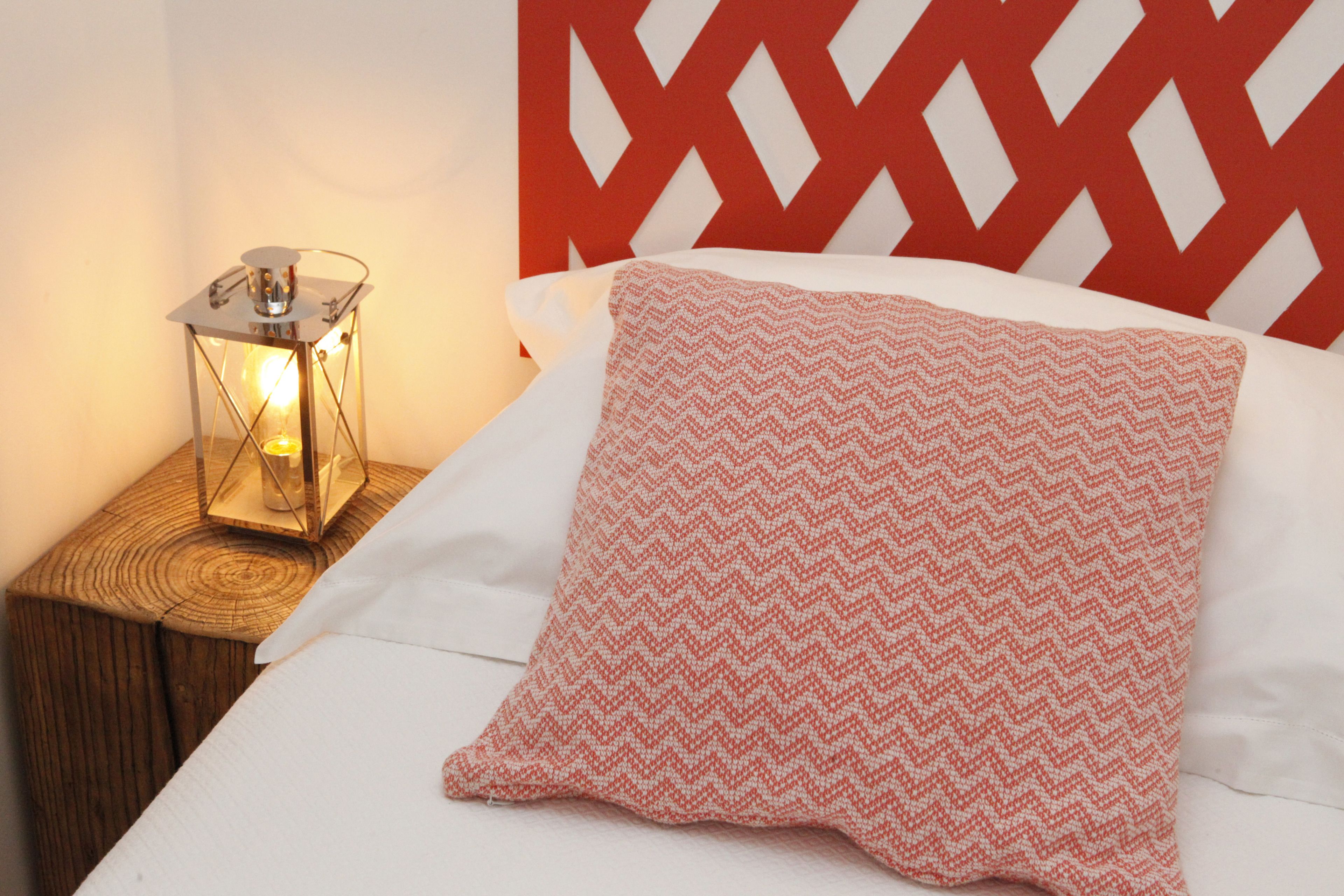 A neatly made bed with a white sheet and a pink and white patterned pillow, a wooden nightstand with a lantern, and a red geometric headboard.