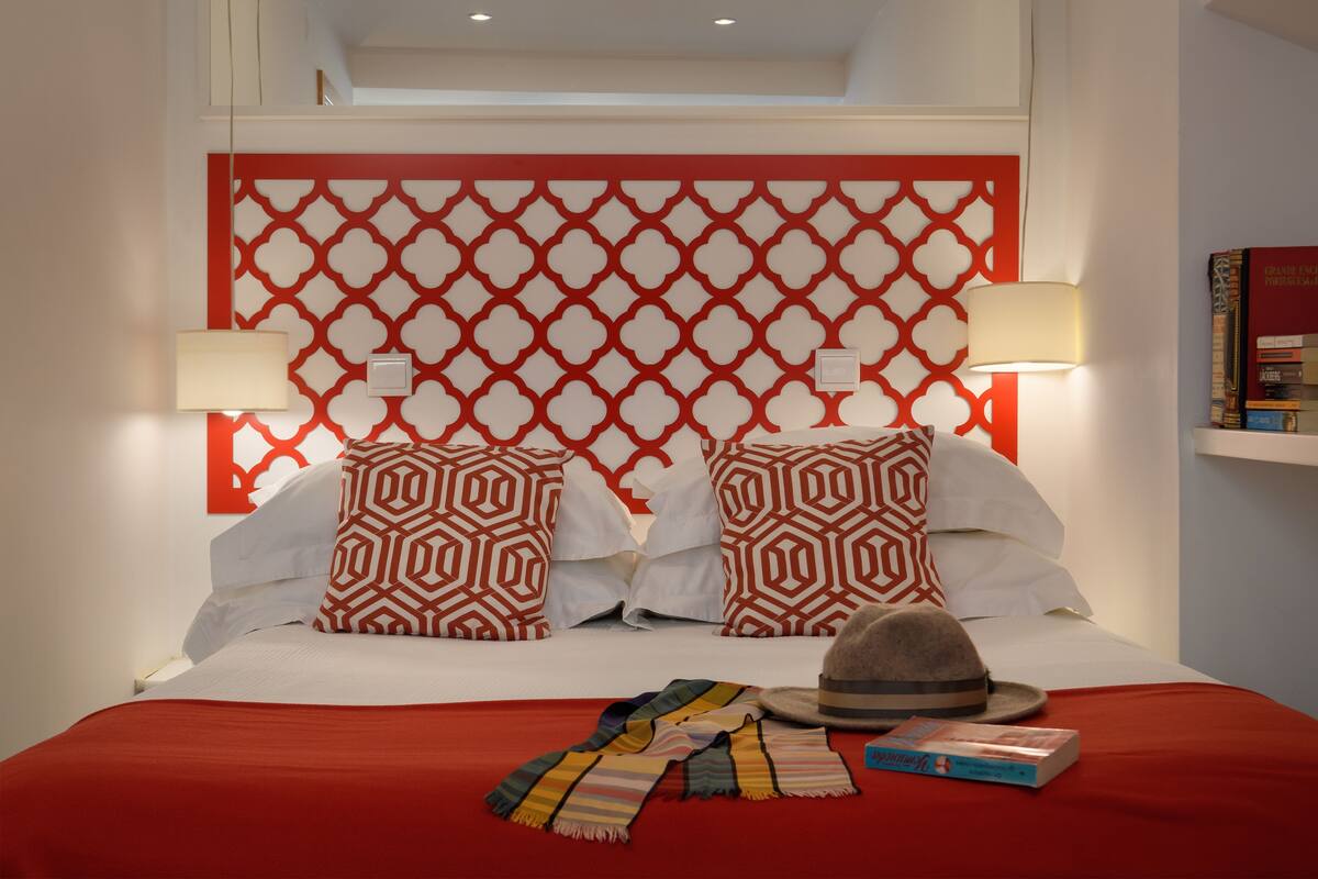 A neatly made bed with patterned pillows, a red blanket, and a hat on the bed. There are two bedside lamps and a book on the bed.
