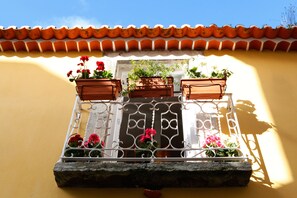 A balcony with terracotta pots containing red flowers and green plants, a white metal railing, and a yellow wall with a tiled roof.