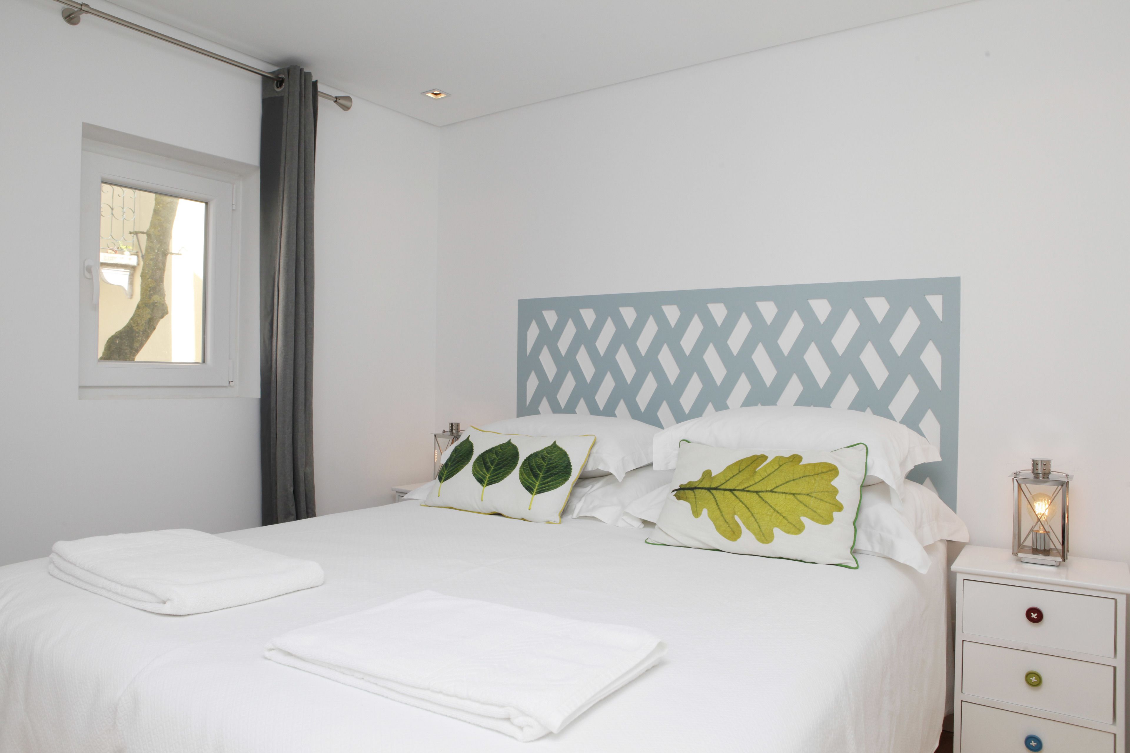 Una cama bien hecha con sábanas y almohadones blancos, una mesita de noche con lámpara, y una ventana con vista a un árbol.