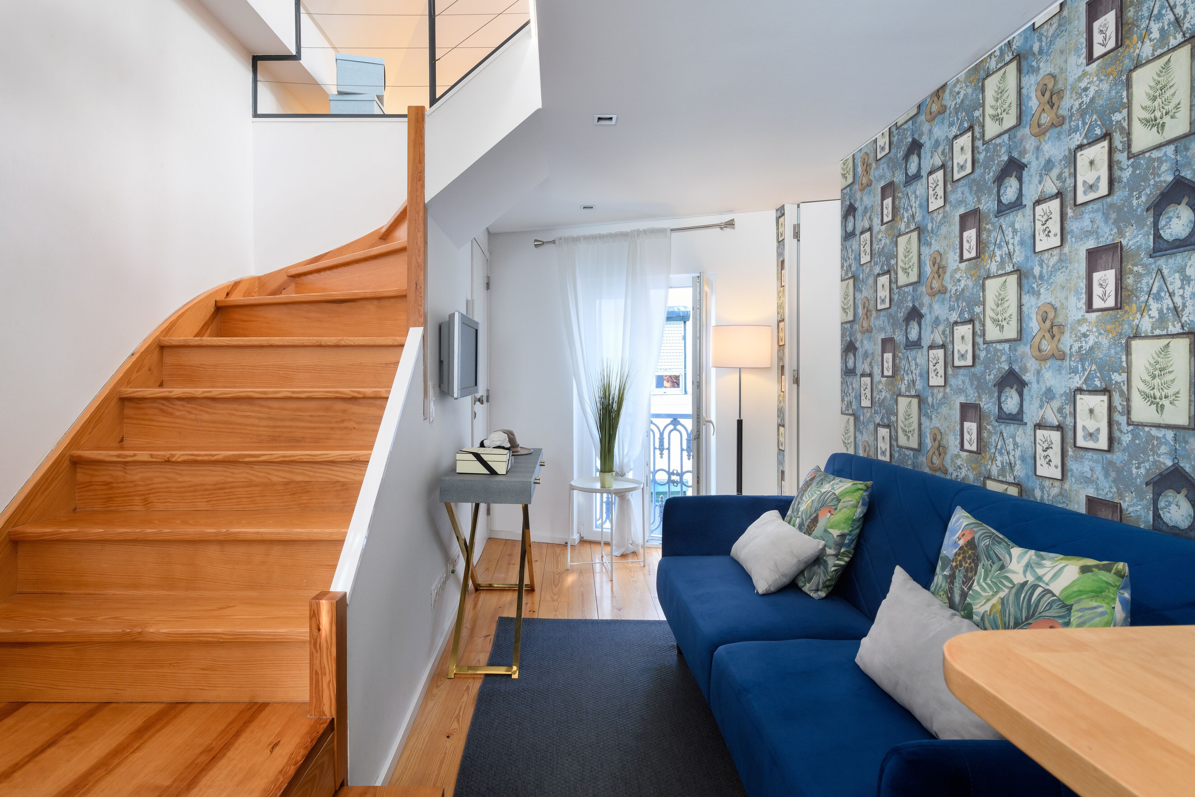 Un salon moderne avec un escalier en bois, un canapé bleu et un mur décoré d’illustrations botaniques encadrées.