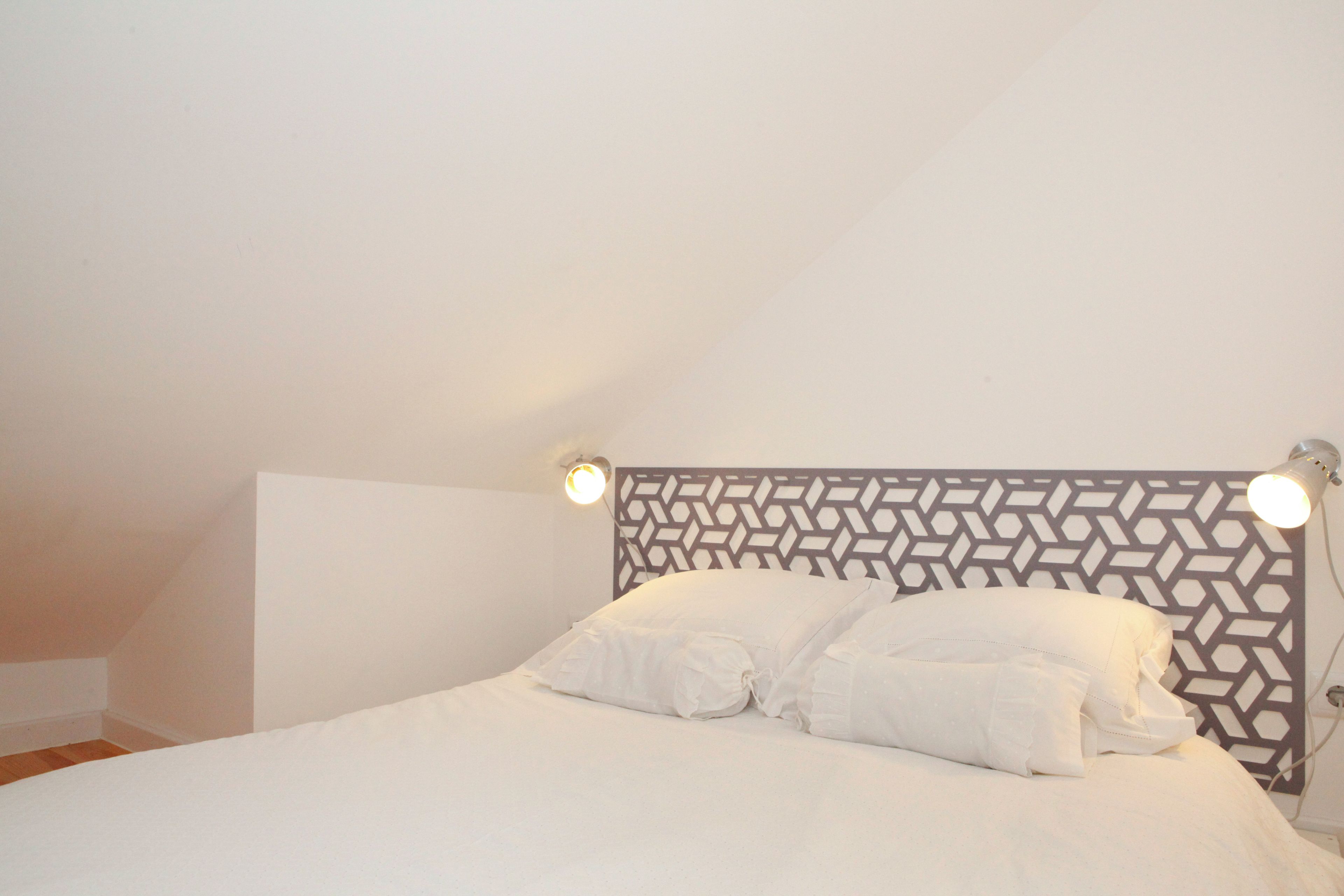 Un dormitorio con una cabecera de diseño geométrico, una cama con ropa de cama blanca y dos lámparas de lectura fijadas a la pared.
