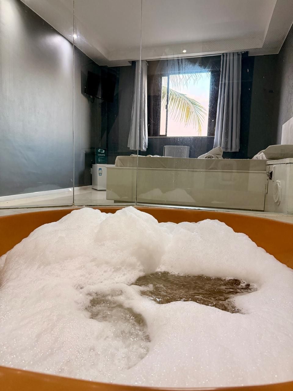 Eine mit Blasen gefüllte Badewanne, ein Fenster mit Vorhängen und ein Palmspiegelbild.