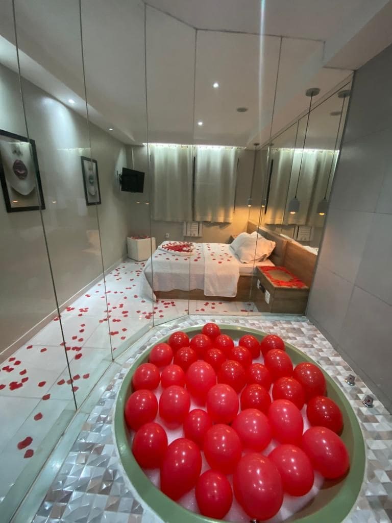 A modern hotel room with a glass partition, a bed with rose petals, and a bowl of red balloons.