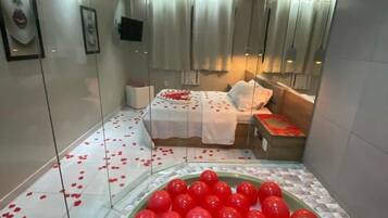 A modern hotel room with a glass partition, a bed with rose petals, and a bowl of red balloons.