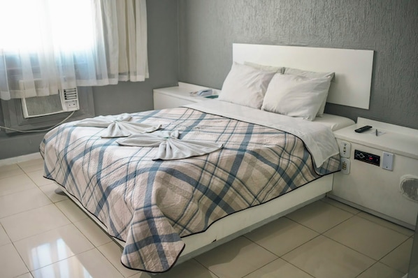 Una cama bien hecha con colcha escocesa, cabecera blanca y mesita de noche con control remoto y toma de corriente.