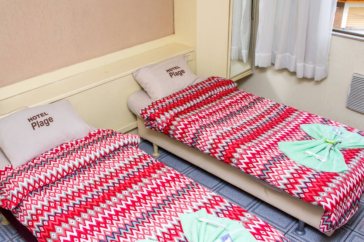 Two single beds with patterned bedding, a window with white curtains, and a wall-mounted mirror.
