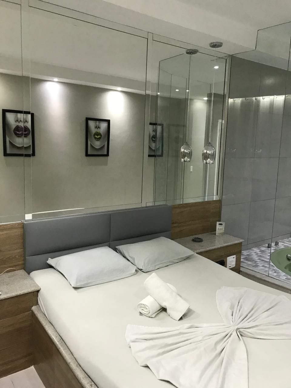 Quarto de hotel moderno com uma cama grande, uma mesinha de cabeceira, um espelho e área de chuveiro.