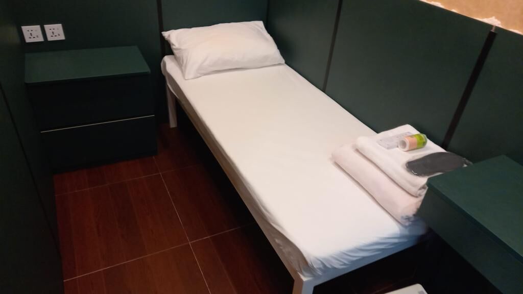 A small, single-bed room with a green headboard, a bedside table, and a power outlet.
