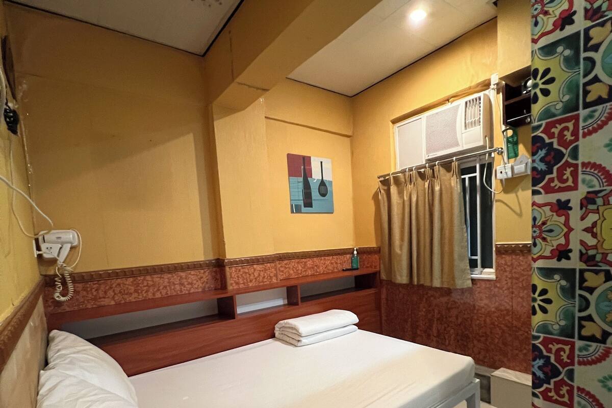 A single bed with a white mattress and pillows, a small bedside table with a bottle, a wall-mounted air conditioner, and a colorful patterned curtain.