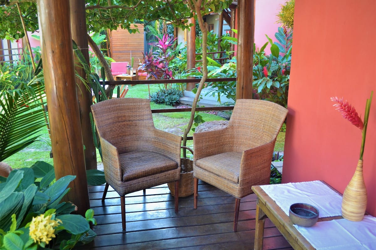 A wooden deck with wicker chairs and a small table, surrounded by lush greenery and a pink wall.