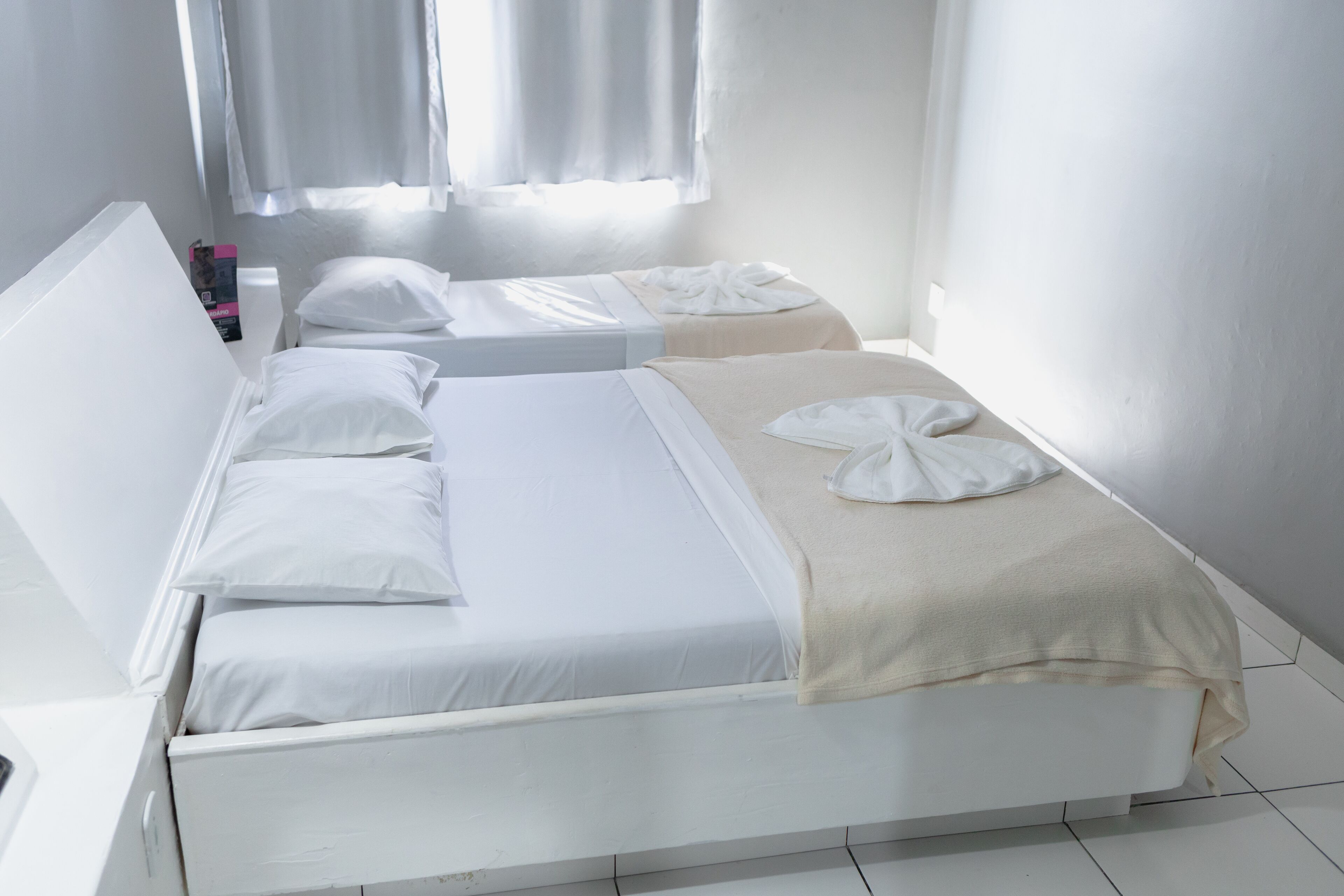 Quarto com duas camas, roupa de cama branca e janela com cortinas.