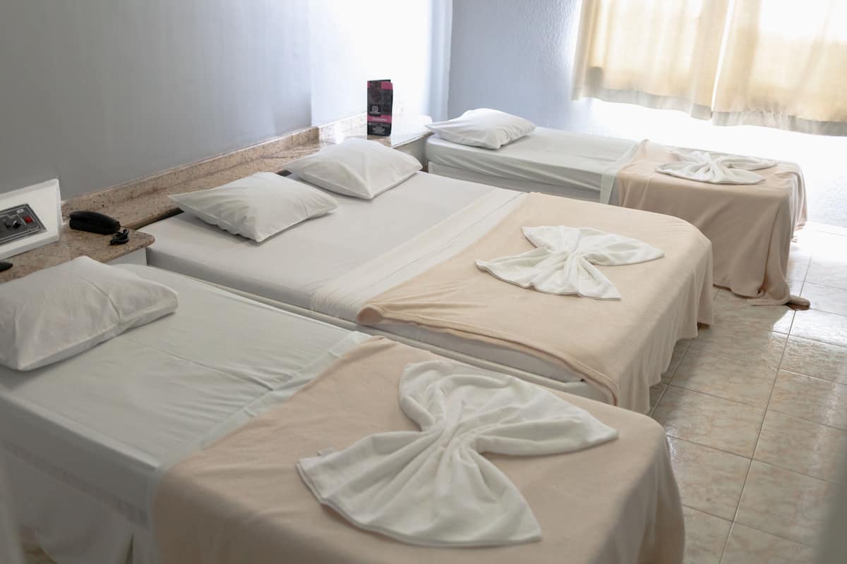 A hotel room with three beds, each with white linens and a towel on the bed. There is a small table with a phone and a box on it.
