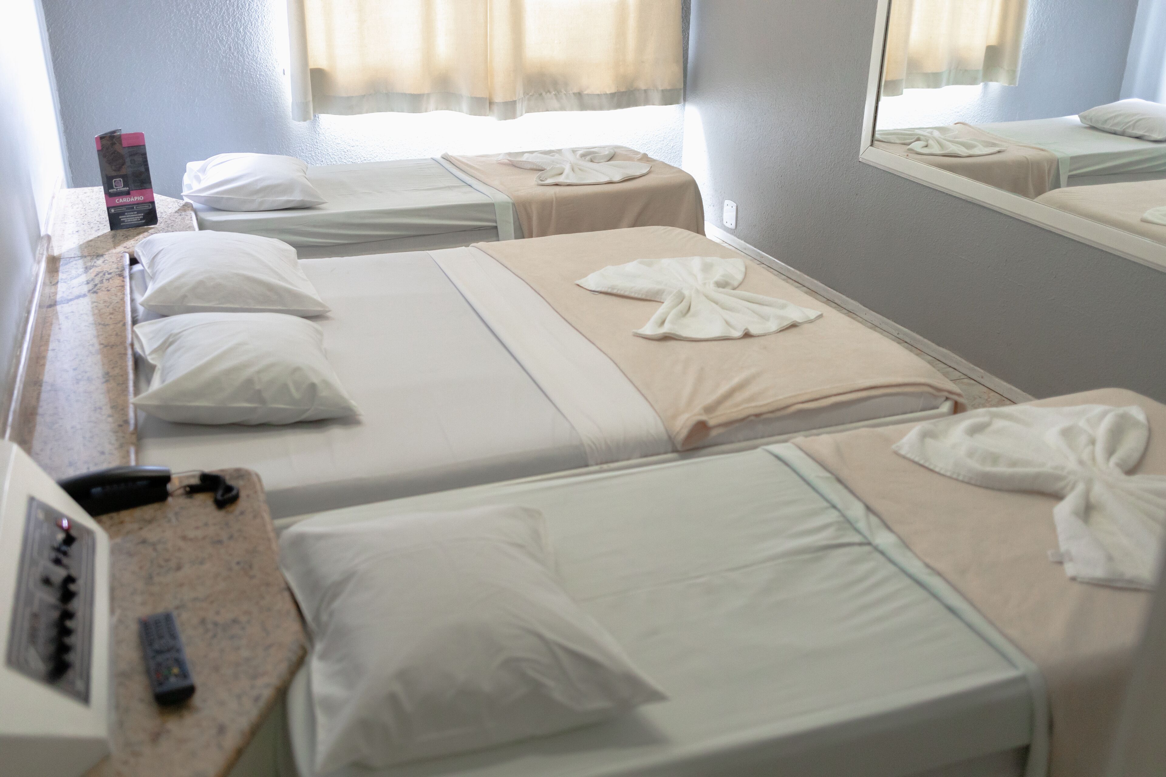 A hotel room with two beds, each with white linens and towels arranged in a bow. A minibar and a control panel are visible on the dresser.