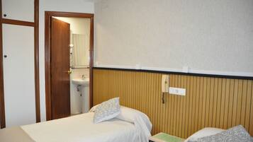 A hotel room with two beds, a wooden headboard, a bedside table with a lamp, and a bathroom visible through an open door.
