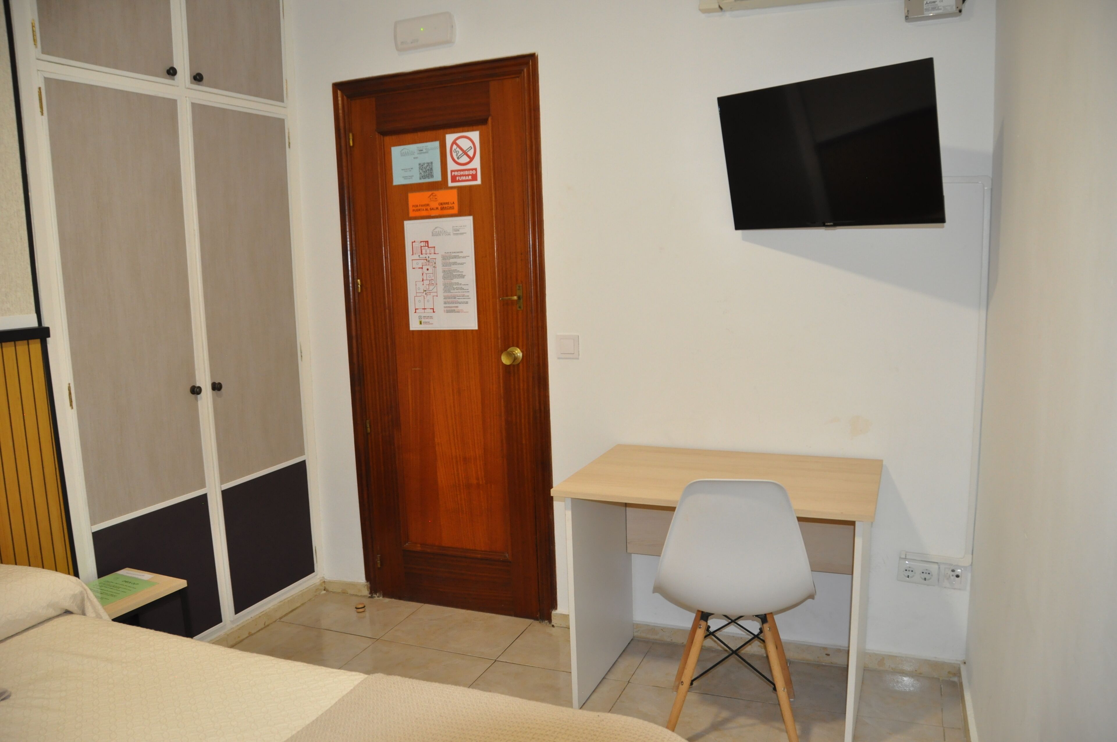 Ein Zimmer mit Holztür, Schreibtisch mit Stuhl und einem an der Wand befestigten Flachbildfernseher.