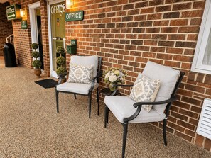 A brick building with an office sign, two chairs with cushions, a small table with a vase, and a key box. - Alleghany Inn (Sparta)