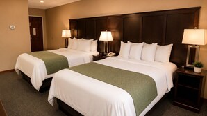 A hotel room with two beds, each with white bedding and a green throw, a dark wood headboard, bedside lamps, a telephone, and a small plant on the nightstand.