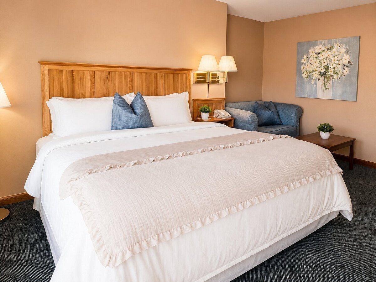 A hotel room with a large bed, two bedside tables, a nightstand, a lamp, a painting, and a carpeted floor.