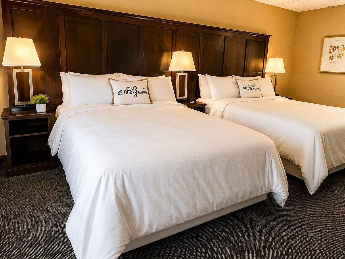 A hotel room with two beds, each with a pillow that says 'Be Our Guest.' There are bedside tables with lamps and a small plant on one of them.