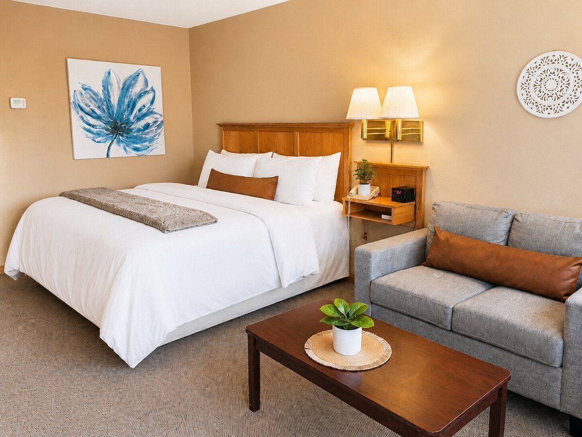 A hotel room with a bed, a sofa, a nightstand with lamps, a wall clock, and a painting.