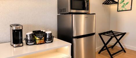 A kitchenette with a coffee maker, a microwave, a refrigerator, and a small table with bags and a basket.