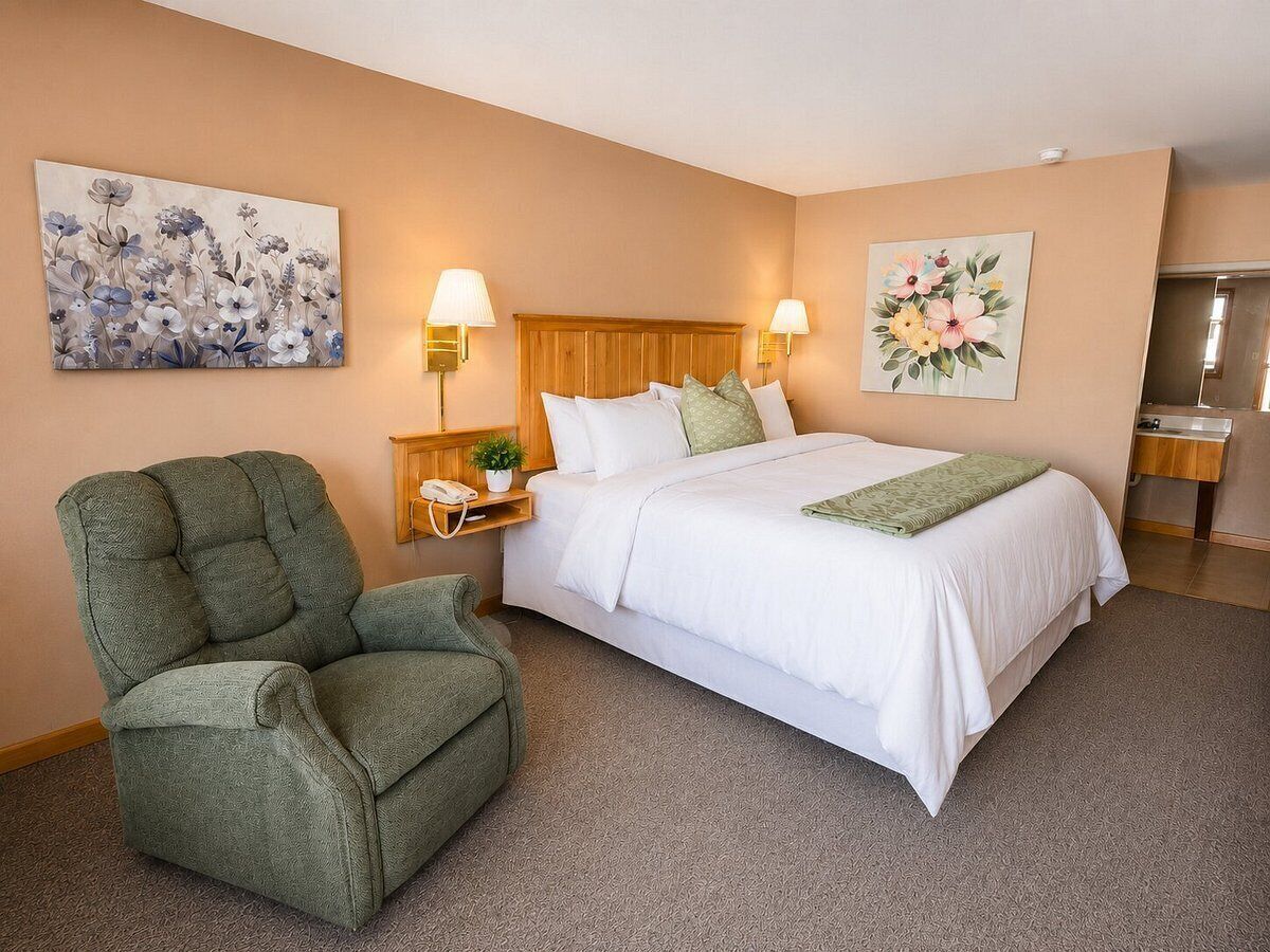 Una habitación de hotel con una cama, un sillón verde, dos lámparas de noche y un cuadro en la pared.