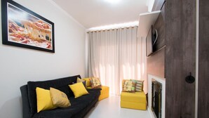 Una sala de estar con un sofá negro, cojines amarillos, un cuadro enmarcado en la pared, chimenea y televisor.
