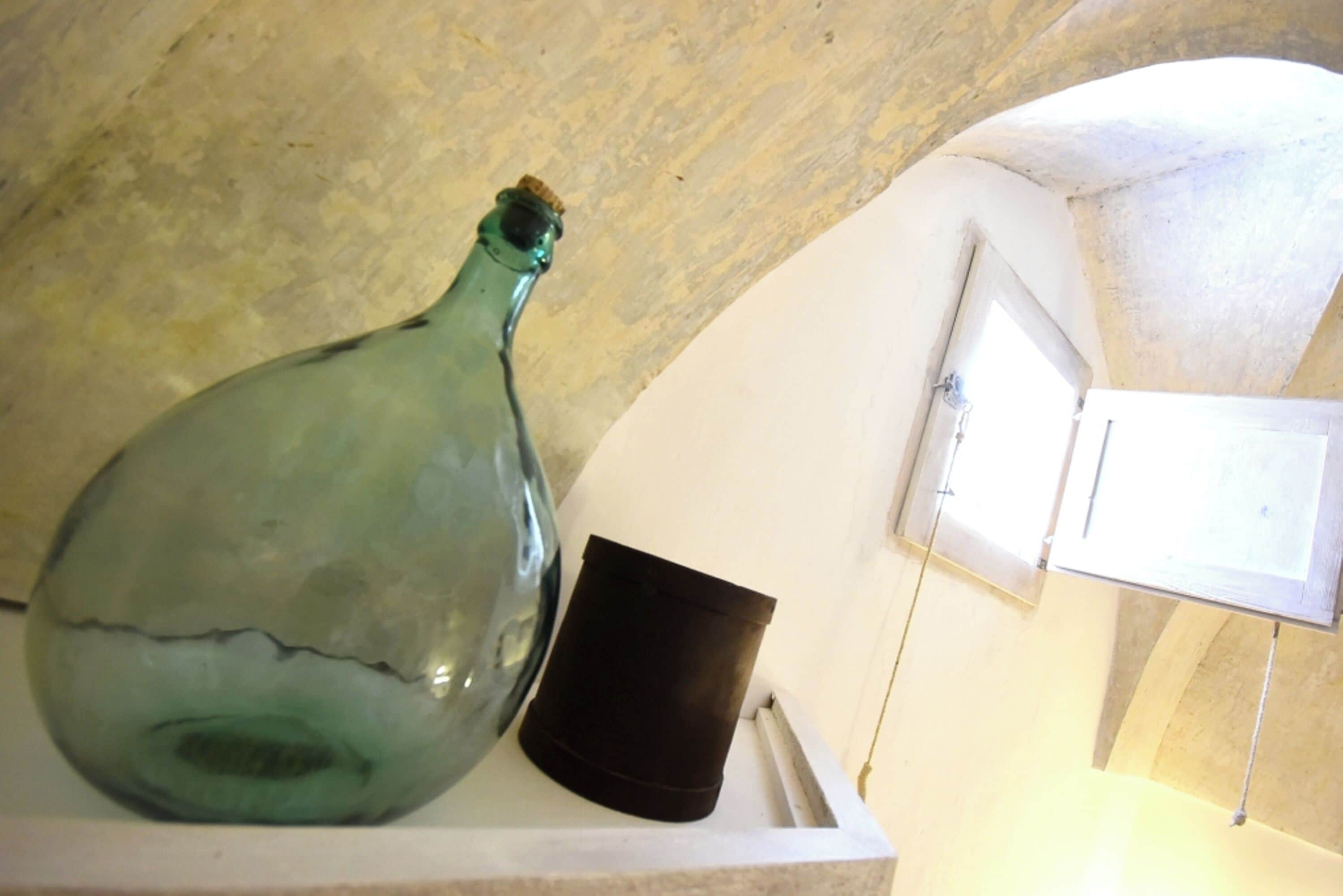 A green glass bottle and a black container on a white shelf against a textured wall.