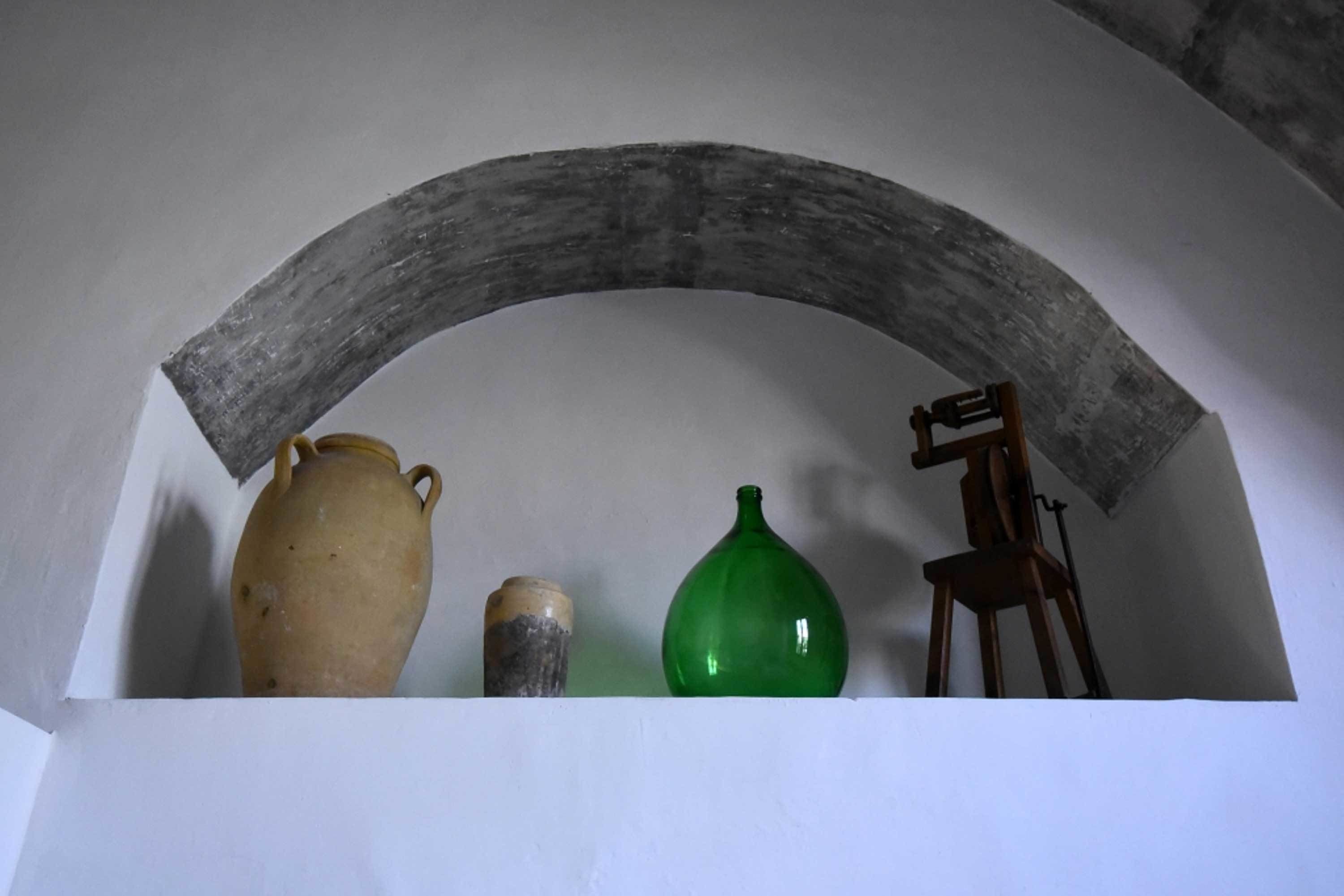 Um quarto com parede branca, arco de madeira, um pote de cerâmica, garrafa verde e um cavalete de madeira.