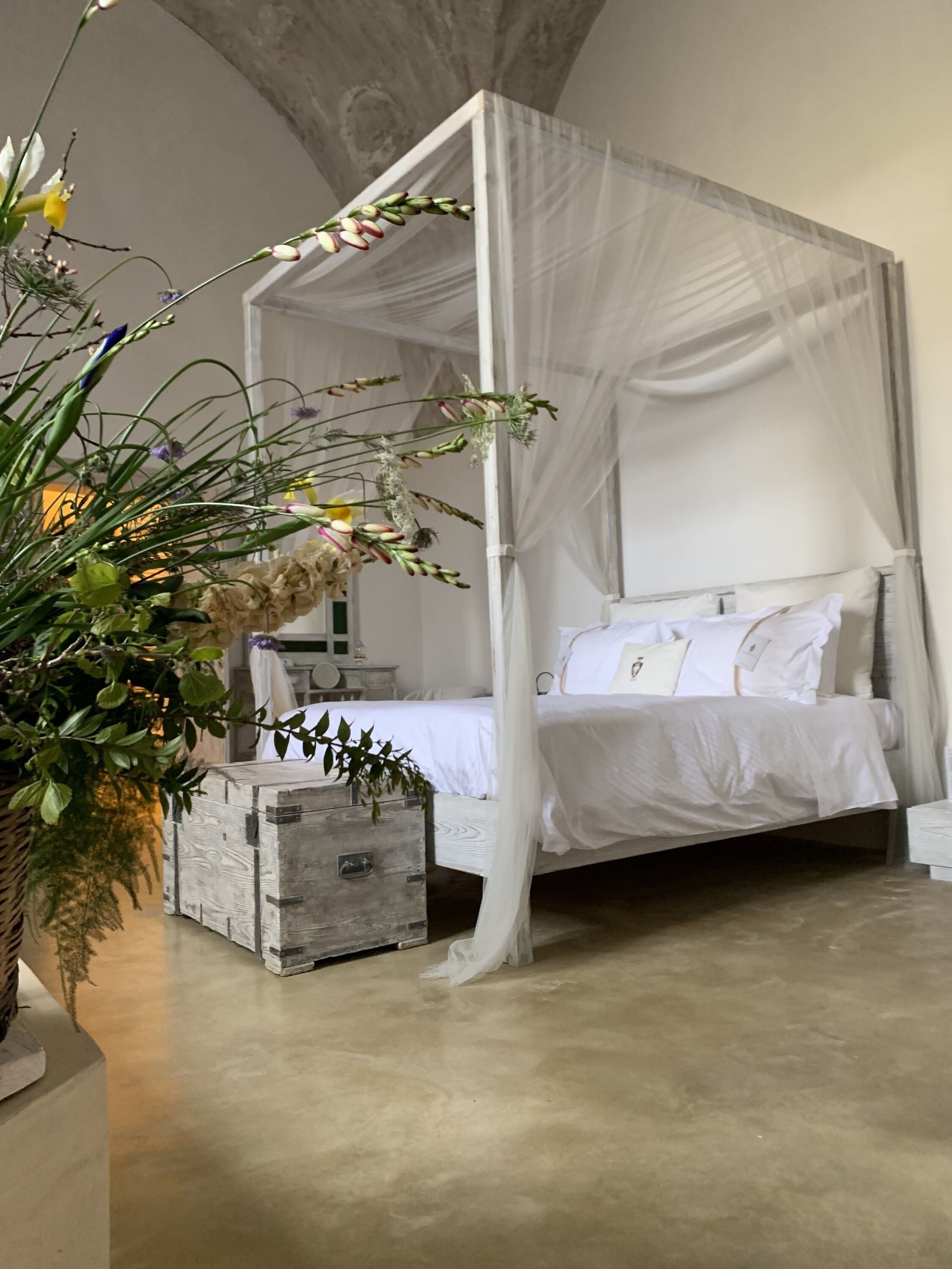 A four-poster canopy bed with white bedding, a wooden chest of drawers, and a large potted plant with green leaves and yellow flowers.