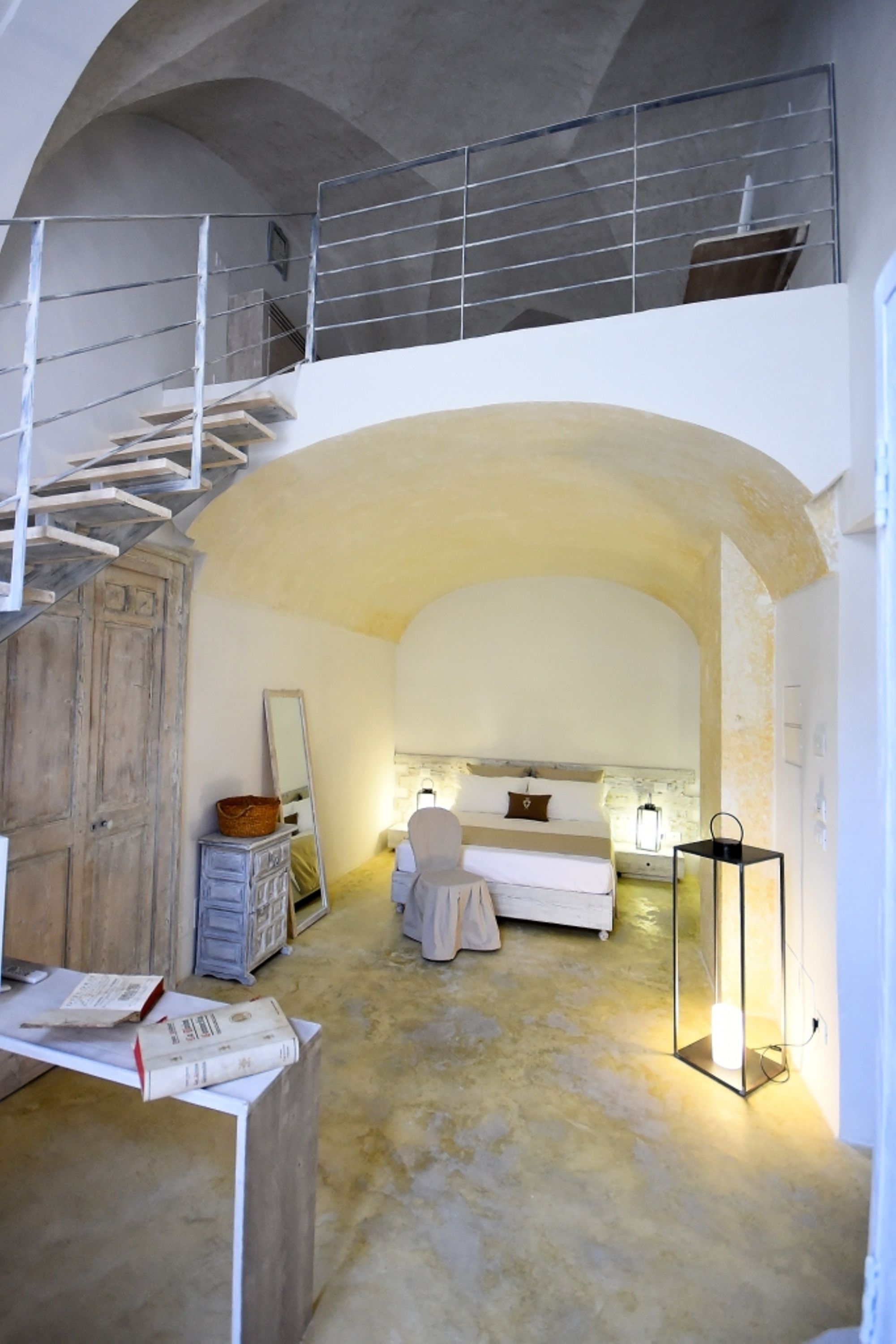 A bedroom with a bed, a desk with books, a mirror, and a staircase leading to an upper level.