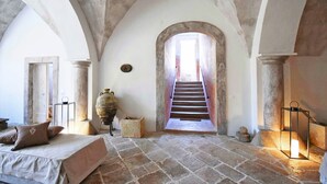 A room with a stone floor, a bed with pillows, a large vase, a lantern, and a staircase leading to another room. - Palazzo Castriota (Alezio)