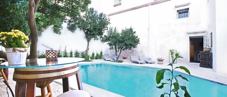 A pool area with a table and chairs, surrounded by greenery and a white building.