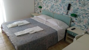 A neatly made bed with white linens and towels, a bedside table with a phone and remote, and a floral wallpaper. - Hotel Corallo (San Mauro Pascoli)