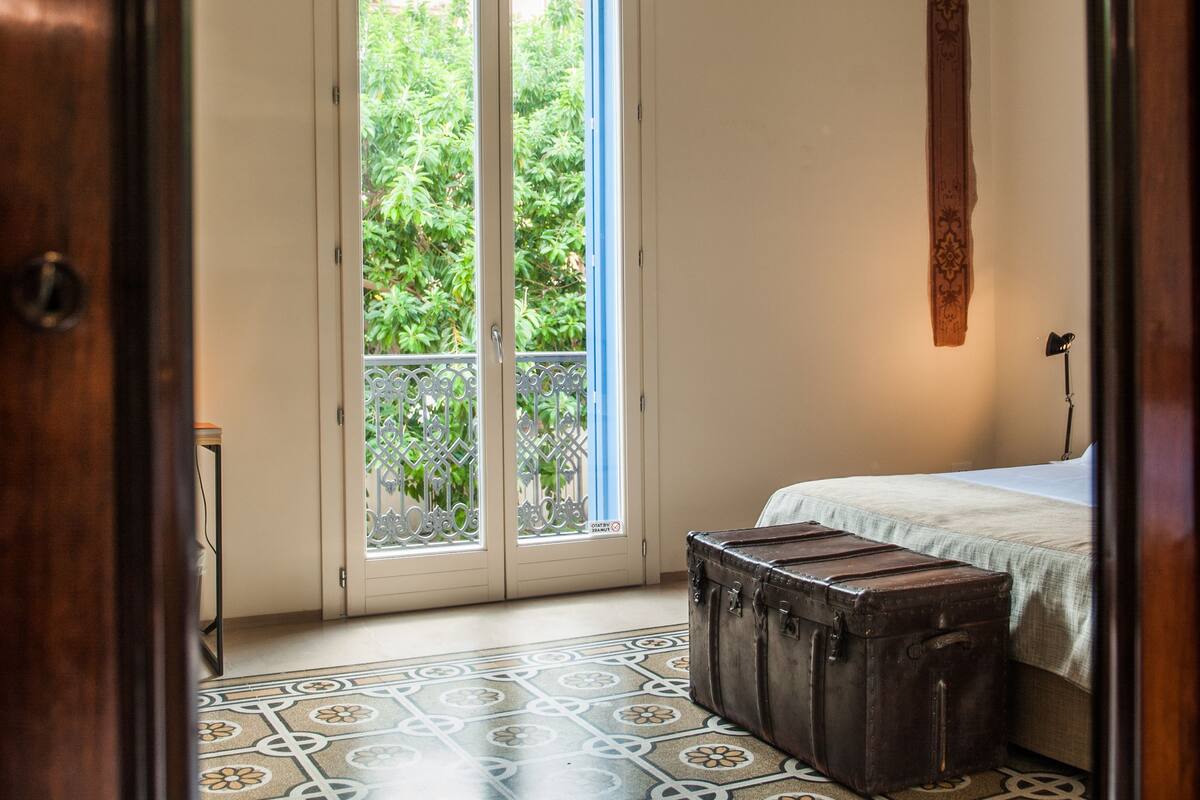 Un dormitorio con un ventanal grande, una cama, un banco de madera y un suelo estampado.