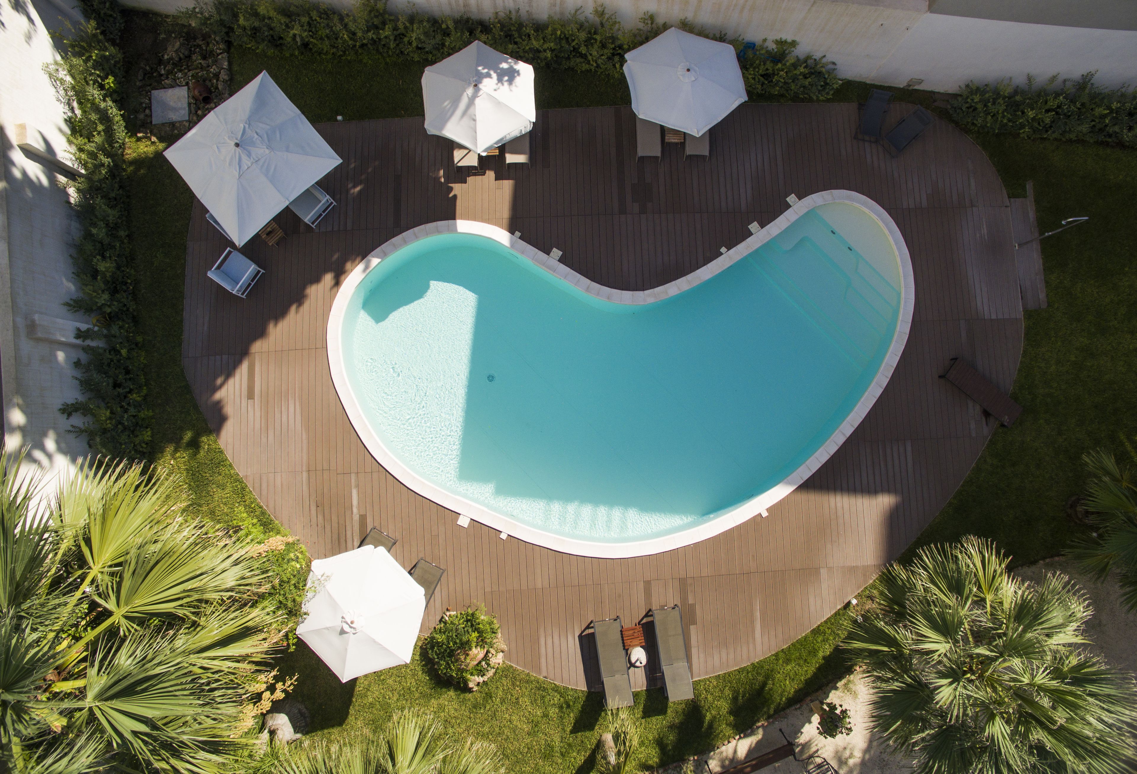 Vue aérienne d’une piscine en forme de cœur, entourée d’une terrasse en bois, de palmiers et de parasols blancs.