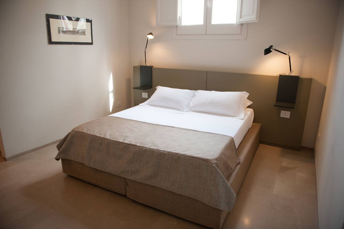 Una cama bien hecha con una colcha color beige, dos lámparas de noche y un cuadro en la pared.