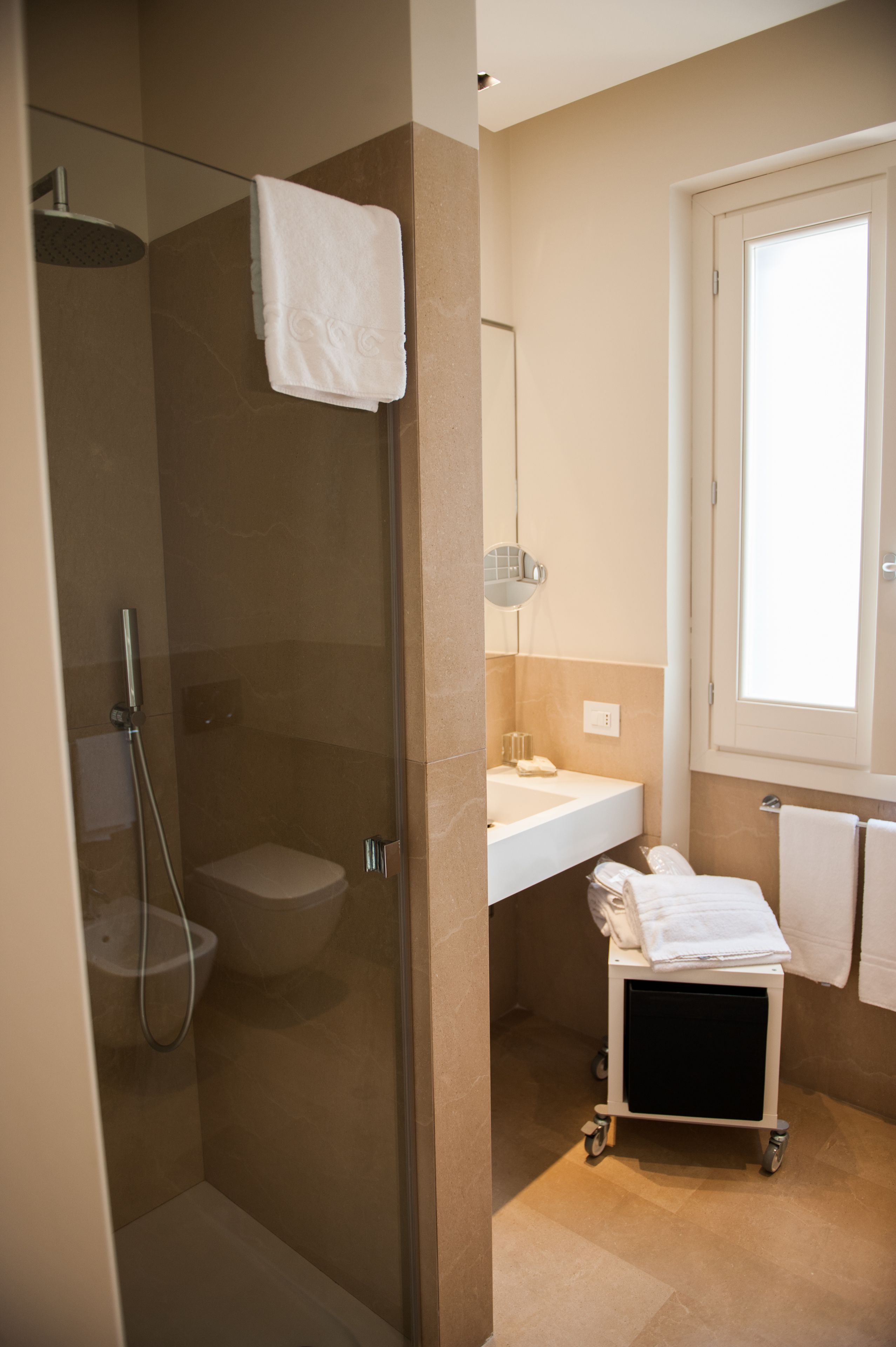 Un baño con ducha de mampara de vidrio, inodoro, lavamanos y carrito con toallas.