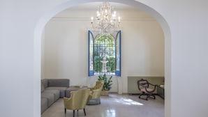 A room with a large arched doorway, a chandelier, a grey sofa, two armchairs, a dining table with chairs, and a stained glass window. - Giardini Mon Plaisir (Trapani)