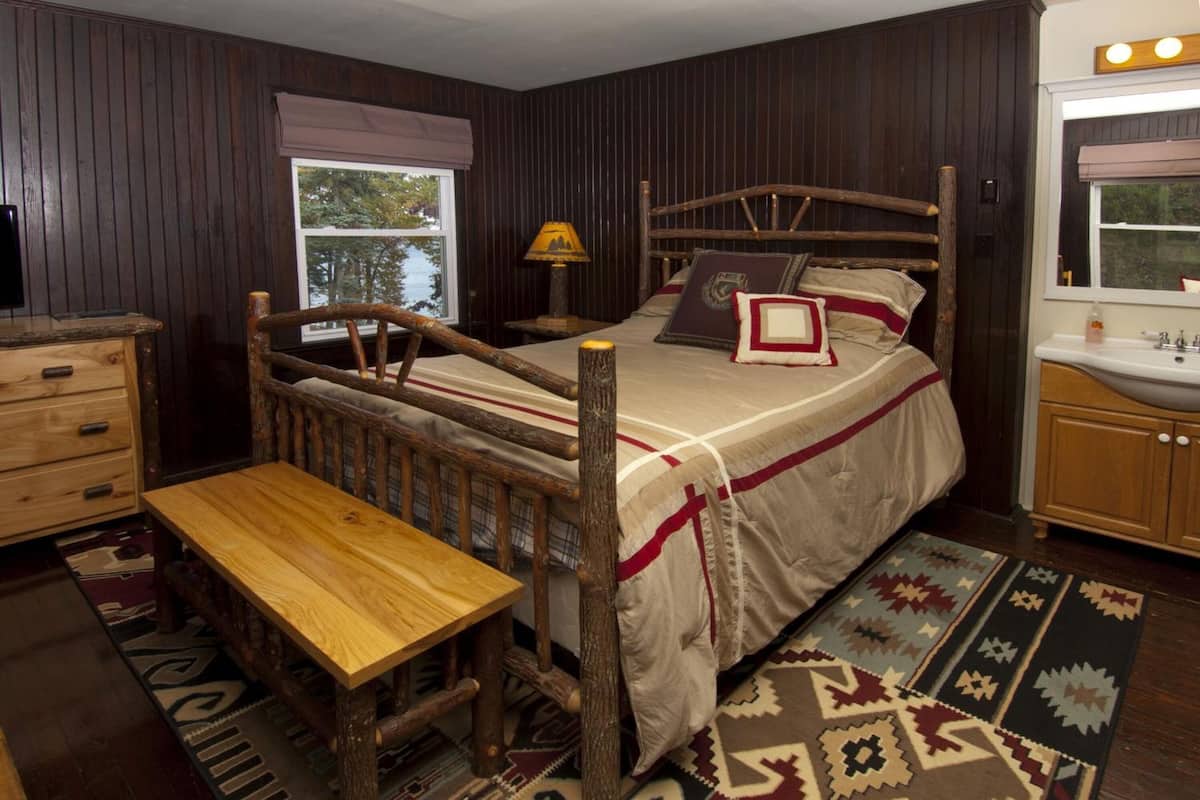 A rustic bedroom with a wooden bed, a wooden bench, a dresser, a window with a view, and a bathroom with a sink and mirror.