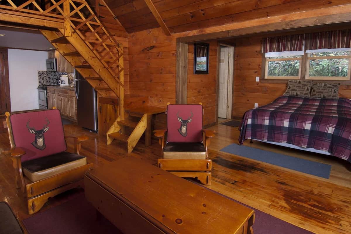 A cabin interior with a wooden staircase, a bed with plaid bedding, and two chairs with deer head cushions.