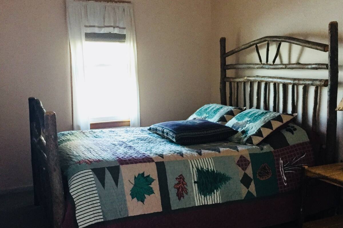 A bed with a patterned bedspread and pillows, a wooden headboard, a window with curtains, and a rug on the floor.
