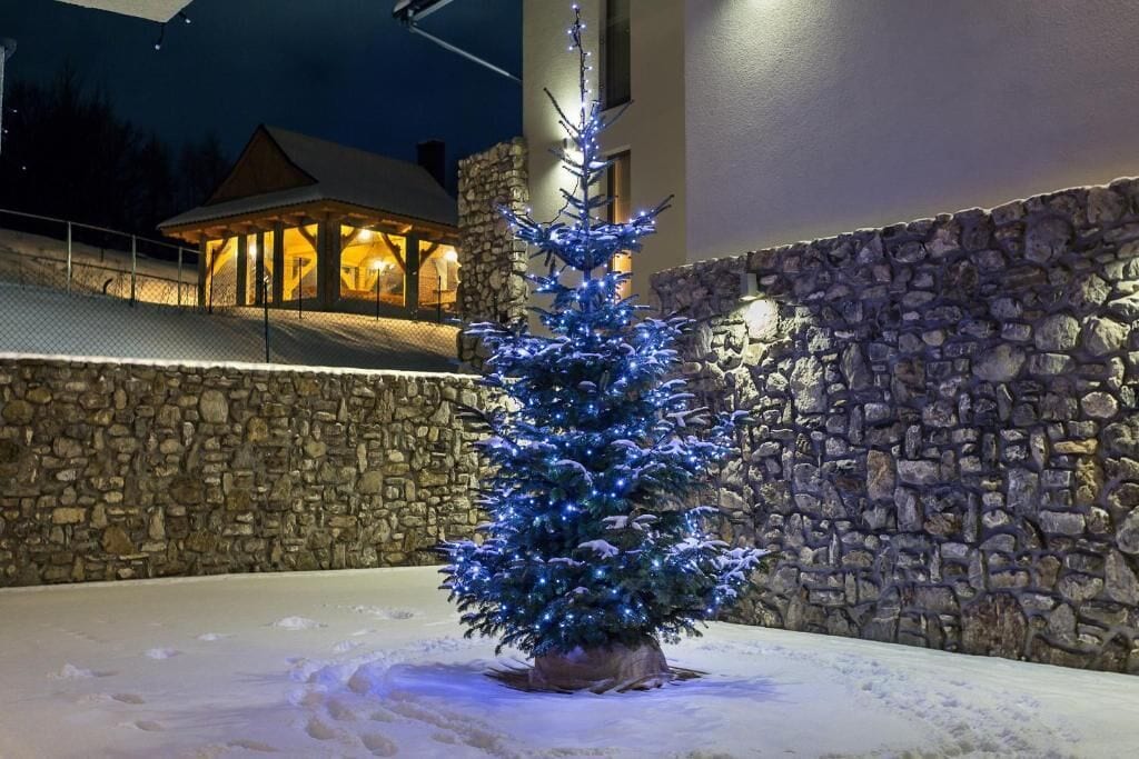 A snow-covered outdoor area with a lit Christmas tree, stone walls, and a building with a wooden structure in the background.